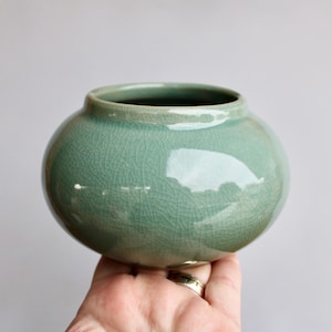 May include: A small, round, celadon-glazed ceramic vase. The vase has a crackle finish and a wide opening. The color is a soft, muted green. The vase is held in a hand.