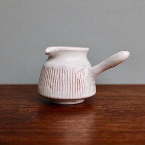 May include: White ceramic creamer with a pink stripe design and a handle. The creamer is sitting on a wooden surface.