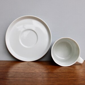 Może przedstawiać: Biały ceramiczny zestaw filiżanki i spodka. Filiżanka ma uchwyt i znajduje się obok spodka. Zestaw znajduje się na drewnianej powierzchni z szarym tłem. Filiżanka i spodek są okrągłe.