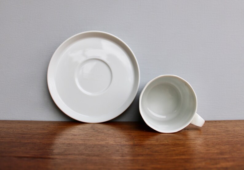 Może przedstawiać: Biały ceramiczny zestaw filiżanki i spodka. Filiżanka ma uchwyt i znajduje się obok spodka. Zestaw znajduje się na drewnianej powierzchni z szarym tłem. Filiżanka i spodek są okrągłe.
