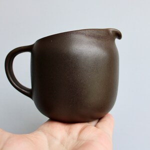 May include: A brown ceramic pitcher with a rounded body and a handle. The pitcher has a smooth, matte finish.
