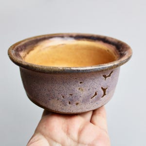 May include: A small, purple and brown ceramic pot with a light brown interior. The pot has a textured surface and a slightly rounded shape.