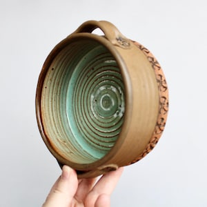May include: A handmade ceramic bowl with a brown exterior and a turquoise interior. The bowl has a handle and a decorative floral pattern around the rim. The bowl is approximately 6 inches in diameter.