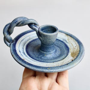 May include: A blue and white ceramic candle holder with a twisted handle. The holder has a circular base and a raised center for the candle.
