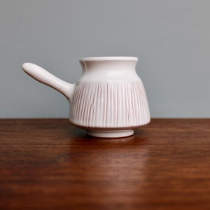 May include: A white ceramic creamer with a pink striped design and a handle. The creamer is sitting on a wooden surface.