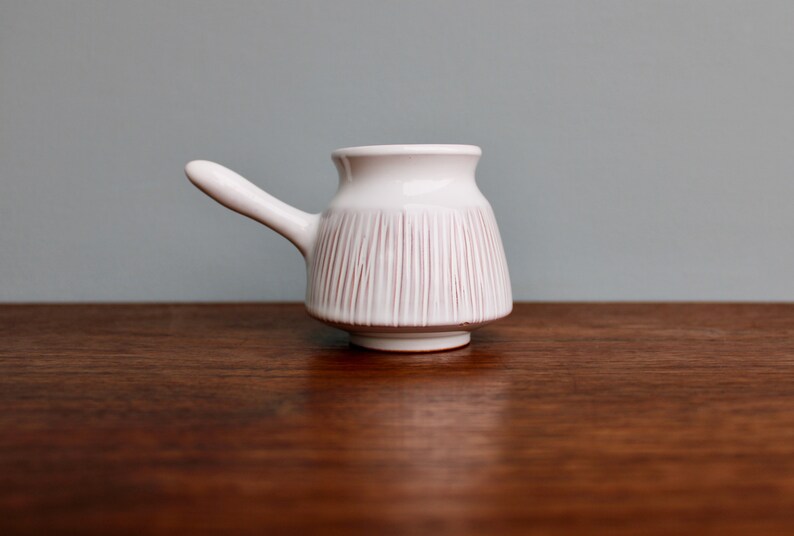 May include: A white ceramic creamer with a pink striped design and a handle. The creamer is sitting on a wooden surface.