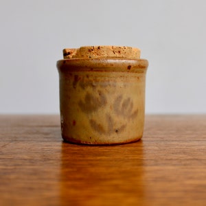 May include: A small, tan ceramic jar with a cork lid. The jar has a floral design in a darker shade of brown.