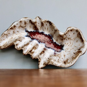 May include: A white ceramic dish with a brown and red glaze. The dish has a wavy, organic shape and a deep, central cavity.