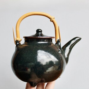 May include: A dark green ceramic teapot with a bamboo handle. The teapot has a rounded body, a spout, and a lid. The handle is arched and attached to the pot with twine. The teapot is likely used for brewing tea.