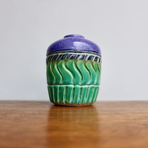 May include: A small, hand-crafted ceramic vase with a purple top and green body. The vase has a wavy, textured design.