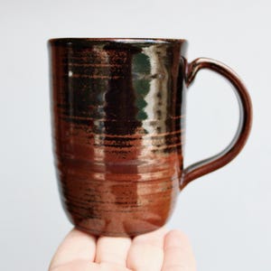 May include: A handmade, brown ceramic mug with a curved handle. The mug has a glossy finish with horizontal, textured lines. The mug is held by a hand against a white background. The mug is approximately 5 inches tall.