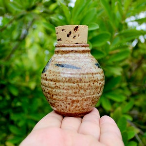 May include: A small, brown and tan ceramic jar with a cork lid. The jar has a speckled glaze and a few blue stripes. The jar is being held in a hand.