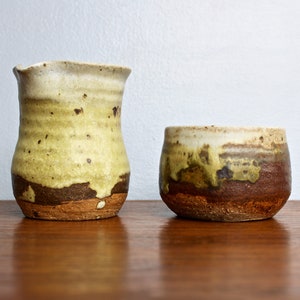 May include: A set of two handmade ceramic pottery pieces. The larger piece is a pitcher with a light brown, yellow, and white glaze. The smaller piece is a cup with a similar glaze. Both pieces have a rustic, handmade look.