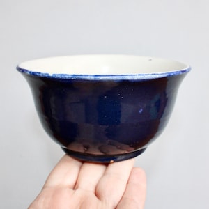 May include: A handmade ceramic bowl with a deep blue exterior and a white interior. The rim is accented with a thin blue line. The bowl has a rounded shape and sits on a small base. The bowl is held by a hand.