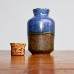 May include: A handmade ceramic vase with a cork stopper. The vase is a mix of blue and brown tones, with a textured surface. The cork stopper is light brown and cylindrical. The vase is on a wooden surface.