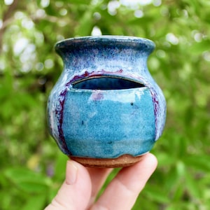 May include: A small, handmade ceramic vase with a unique design. The vase features a vibrant turquoise glaze with streaks of purple and a dark blue rim. It has a distinctive opening on the side, perfect for small plants or decorative items.