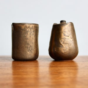 May include: Two small ceramic containers with a metallic gold glaze. The containers have a textured surface and are sitting on a wooden surface.