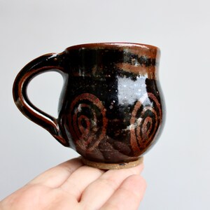 May include: A small, dark brown ceramic mug with a rounded body and a handle. The mug has a black glaze with brown swirls and speckles.