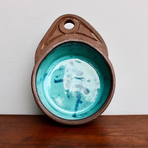 May include: A small, brown ceramic bowl with a turquoise glaze. The bowl has a handle with a hole in the top.