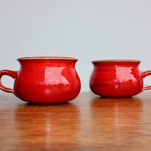 May include: Two red ceramic mugs with a rounded shape and a single handle. The mugs are sitting on a wooden surface.