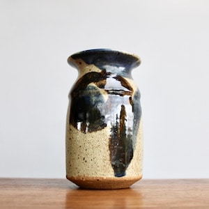 May include: A handmade ceramic vase with a speckled beige body and a dark blue rim. The vase features dark brown and blue glaze drips. The vase is cylindrical with a slightly flared top, perfect for displaying flowers or as a decorative piece.