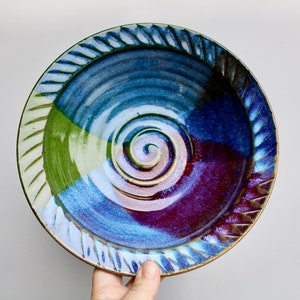 May include: Handmade ceramic plate with a spiral design in the center. The plate features a color palette of blue, green, and purple. The rim has a textured, ridged pattern. A unique piece for serving or display.