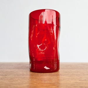 May include: A vibrant red glass tumbler with a textured, organic shape. The translucent glass catches the light, showcasing the rich color. The tumbler sits on a wooden surface against a white background.