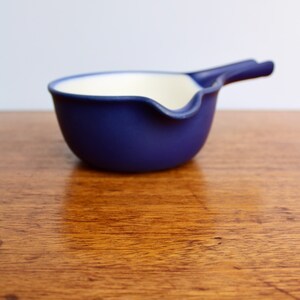 May include: A small, blue ceramic dipping bowl with a white interior and a curved handle. The bowl is sitting on a wooden surface.
