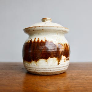 May include: A white ceramic jar with a lid, decorated with brown glaze in a drip pattern. The jar has a rounded body and a slightly flared rim.