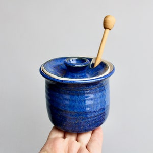 May include: A blue ceramic honey pot with a wooden honey dipper. The pot has a lid with a small hole for the dipper.