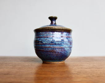 May include: A small, blue and brown ceramic jar with a lid. The jar has a mottled glaze with streaks of purple and white.