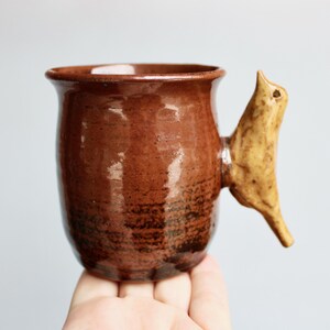 May include: A glossy, dark brown ceramic mug with a smooth, rounded body and a flared rim. The handle is uniquely shaped like a light tan, speckled bird, extending outward from the side of the mug, providing a distinct and tactile grip.