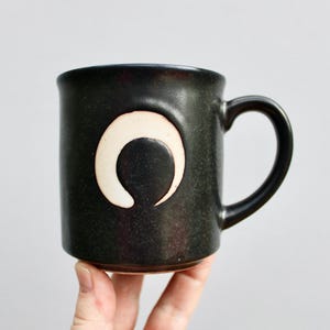 May include: A dark green ceramic mug with a white crescent moon design. The mug has a rounded handle.