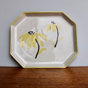 May include: A white ceramic octagonal tray with a gold rim, featuring two yellow flowers with black centers painted on the surface.