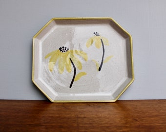 May include: A white ceramic octagonal tray with a gold rim, featuring two yellow flowers with black centers painted on the surface.