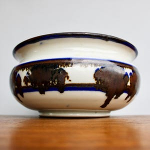 May include: A white ceramic bowl with a brown and blue design. The bowl has a rounded shape and a wide rim. The design features abstract shapes and lines in brown and blue.