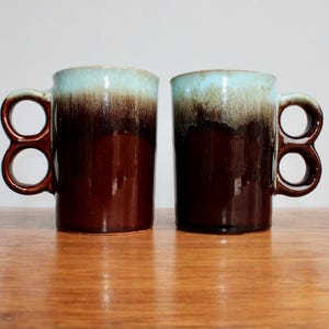 May include: Two brown and blue ceramic mugs with double handles. The mugs have a glazed finish and a light blue rim.