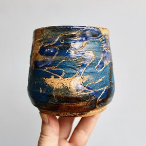 May include: A ceramic cup with a textured, uneven surface featuring a deep blue and brown glaze. The exterior displays a swirling, abstract pattern of dark blue and earthy brown tones with visible cracks and a rustic, hand-crafted finish.