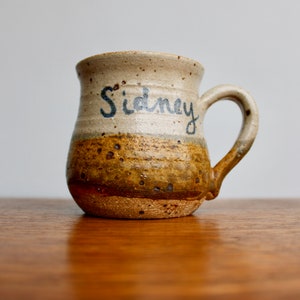 May include: A brown and white ceramic mug with a brown handle. The mug has a brown and white glaze and the name "Sidney" is written on the side in black.