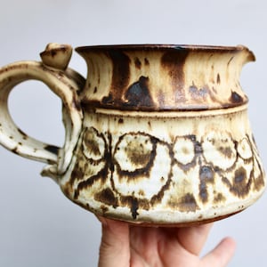 May include: A handmade ceramic pitcher with a unique design. The pitcher features a cream-colored base with brown, organic patterns and a dark brown rim. It has a curved handle and a spout for easy pouring. The pitcher is a decorative and functional piece.