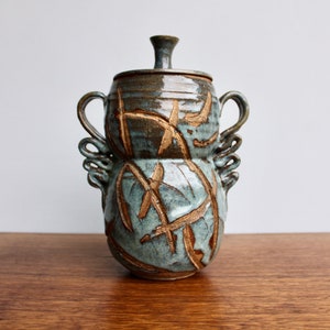 May include: A ceramic jar with a lid and two handles. The jar is decorated with a blue and brown glaze and has a pattern of brown lines and shapes. The jar is sitting on a wooden surface.
