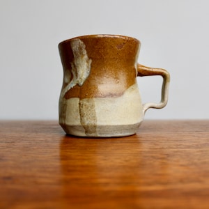 May include: A brown and white ceramic mug with a unique, slightly uneven shape. The mug has a handle and a glazed finish.