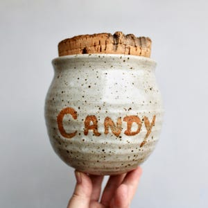 May include: A white ceramic candy jar with a cork lid and the word "CANDY" written in brown glaze.