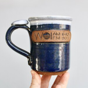 May include: A blue ceramic mug with a brown leather label that reads "WOLI AM 640 FM 90.1".