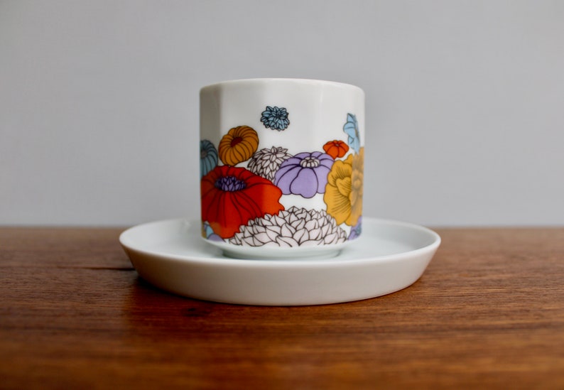 Może przedstawiać: Biały zestaw filiżanki i spodka ceramicznego. Filiżanka ma kolorowy kwiatowy wz&oacute;r z czerwonymi, pomarańczowymi, ż&oacute;łtymi, fioletowymi i niebieskimi kwiatami. Filiżanka stoi na pasującym białym spodku.