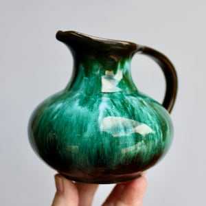 May include: A small green and brown ceramic pitcher with a white glaze. The pitcher has a rounded body and a small handle.