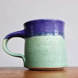 May include: A handmade ceramic mug with a unique color scheme. The top is a deep purple, transitioning to a light green speckled body. The handle is a matching green. This mug is perfect for coffee or tea.