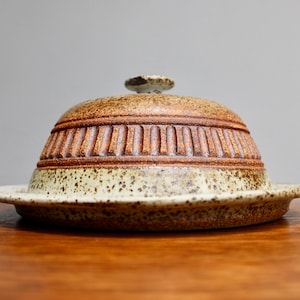 May include: A ceramic butter dish with a speckled brown and white glaze. The lid has a raised, textured design around the edge.