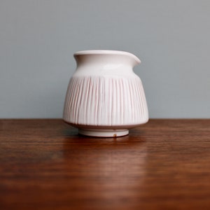 May include: White ceramic creamer with vertical lines, sitting on a wooden surface.
