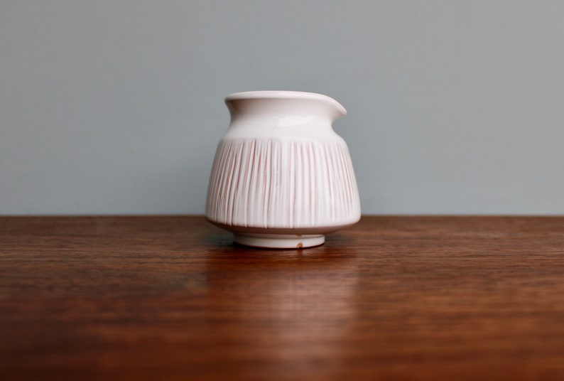 May include: White ceramic creamer with vertical lines, sitting on a wooden surface.